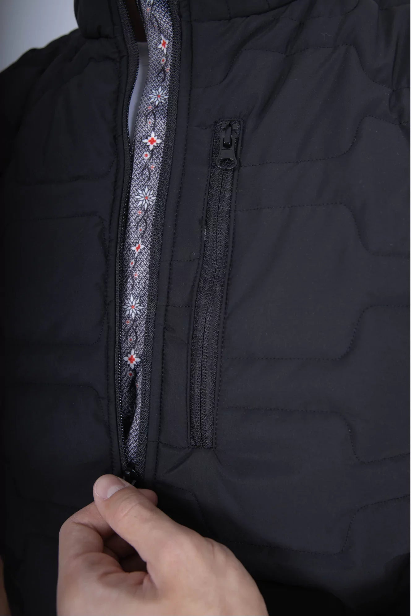 Gilet Edelweiss Herren in Schwarz mit grauem Edelweiss-Muster, hochwertig verarbeitet, mit dezenten Edelweiss-Streifen am Reissverschluss und Nacken, ideal für Arbeit, Freizeit und elegante Anlässe.